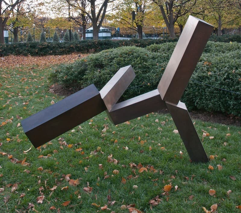 This sculpture features connected geometric forms resembling elongated rectangular blocks positioned at an angle and intersecting in various directions. It is balanced on one thin block on the right, while the other blocks extend in the air out to the left and upward. The colors of the sculpture include dark bronze and metallic brown, and it reflects light coming from above. The sculpture is set in a grassy area with fallen leaves, surrounded by shrubbery and trees with autumnal foliage.